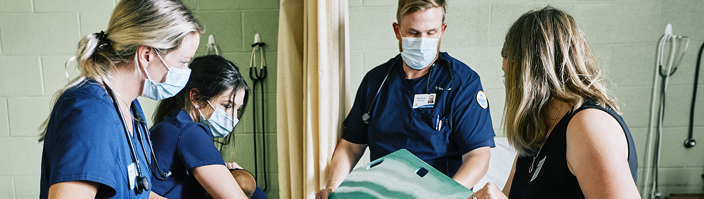 A group of nursing students in PPE gather around a medical bed.