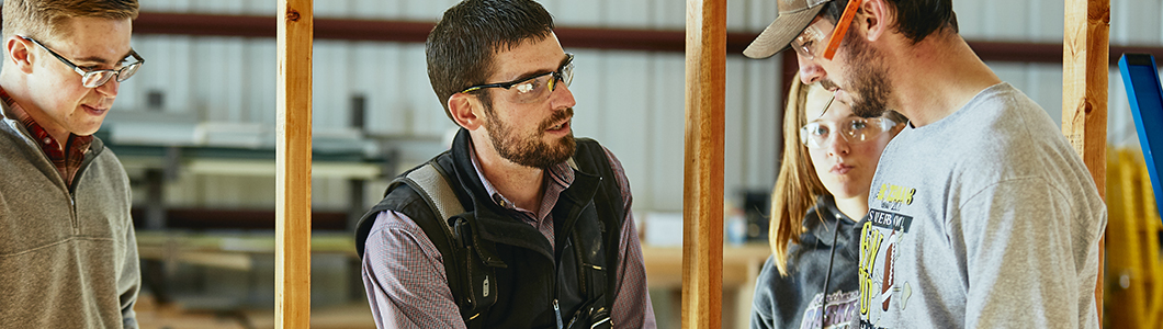 Two men in safety gear talk in a barn while others look on.