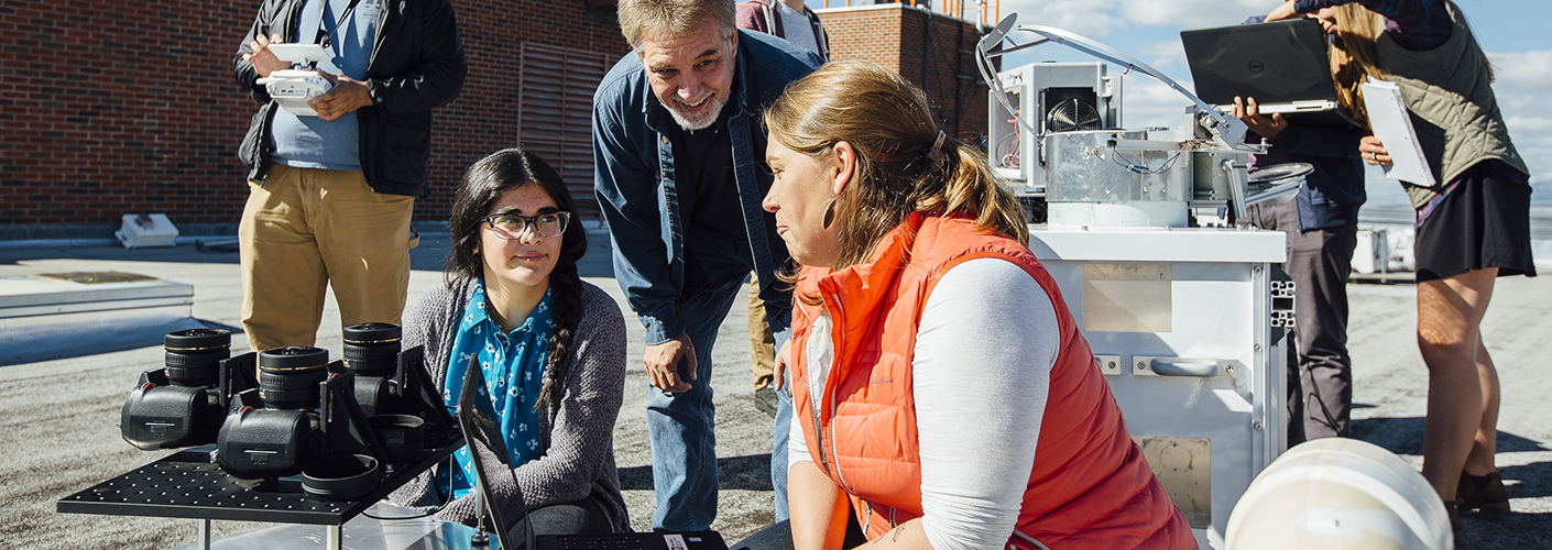 An older man crouches to talk to two young women, seated on the ground, as they inspect scientific instruments.