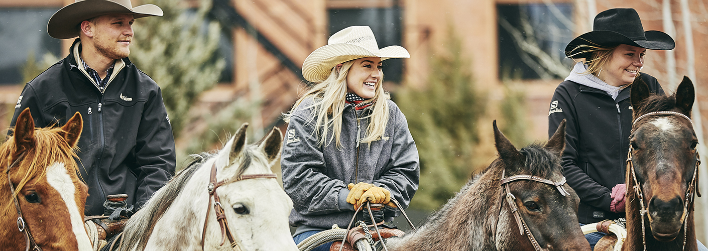 A young man and two young women in cowboy hats sit astride in a line on their horses, laughing and smiling.