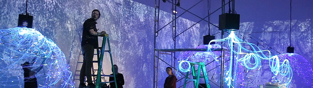 A man stands on a stepladder in a gallery mid-installation of a light-based art display.