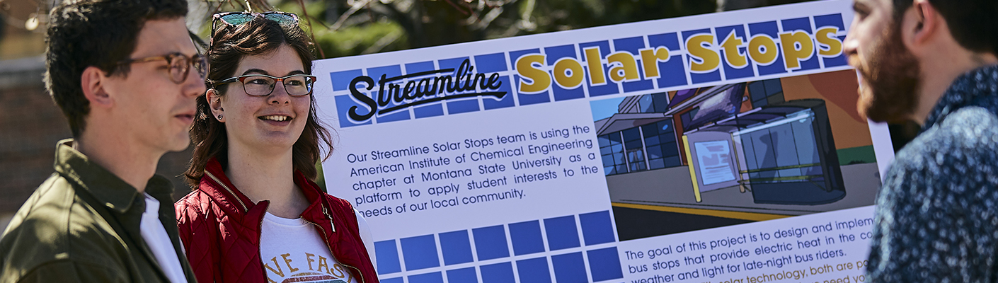 Two students in short sleeves in front of a presentation poster about solar bus stops discuss their project with a bearded man.