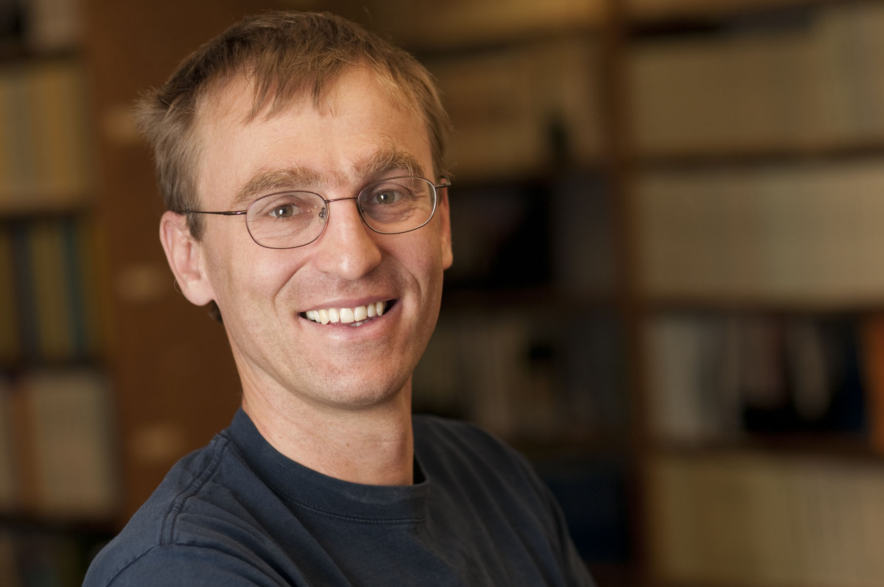 Man with short brown hair and glasses wearing a blue shirt standing in front of a blurred out background of books on a shelf