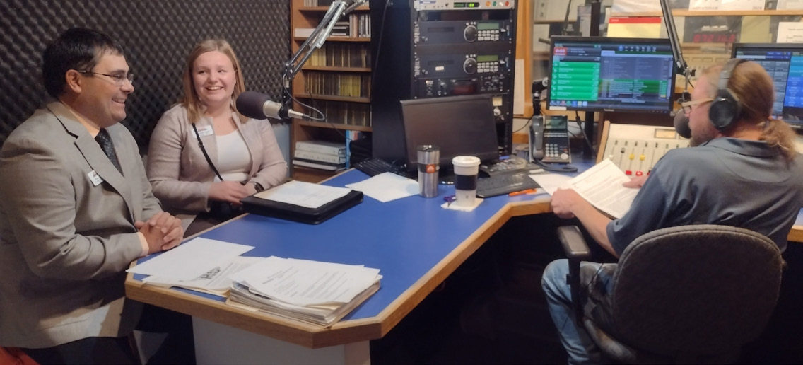 A man and a woman sit in a radio studio, at a table, with a large microphone in between them. The radio host sits on the opposit side of the table.