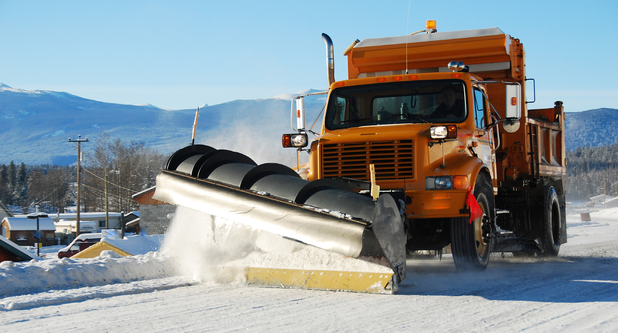 Orange snowplow on a snow covered road, pushing snow to the side with a large black bucket attachment.