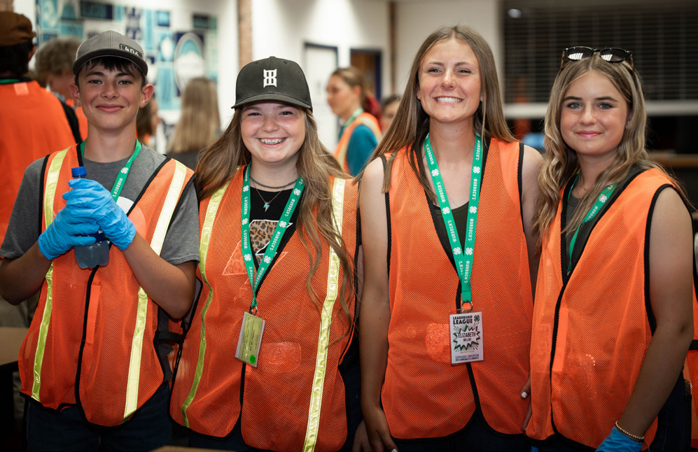 Four young 4-H members stand together, each in a relective orange vest, preparing to engage in an act of hands on community service.