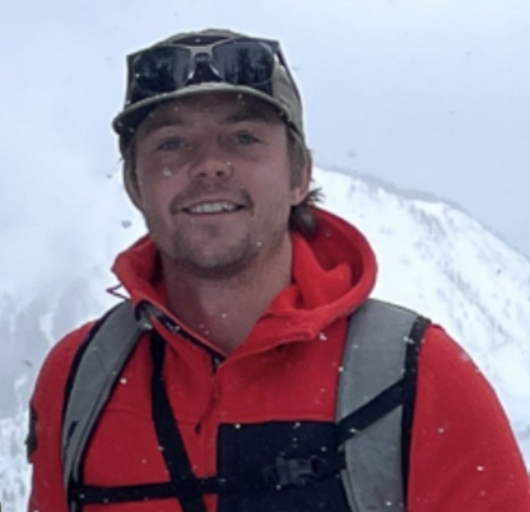 A person wearing a hat and an orange jacket with snowy mountains in the background